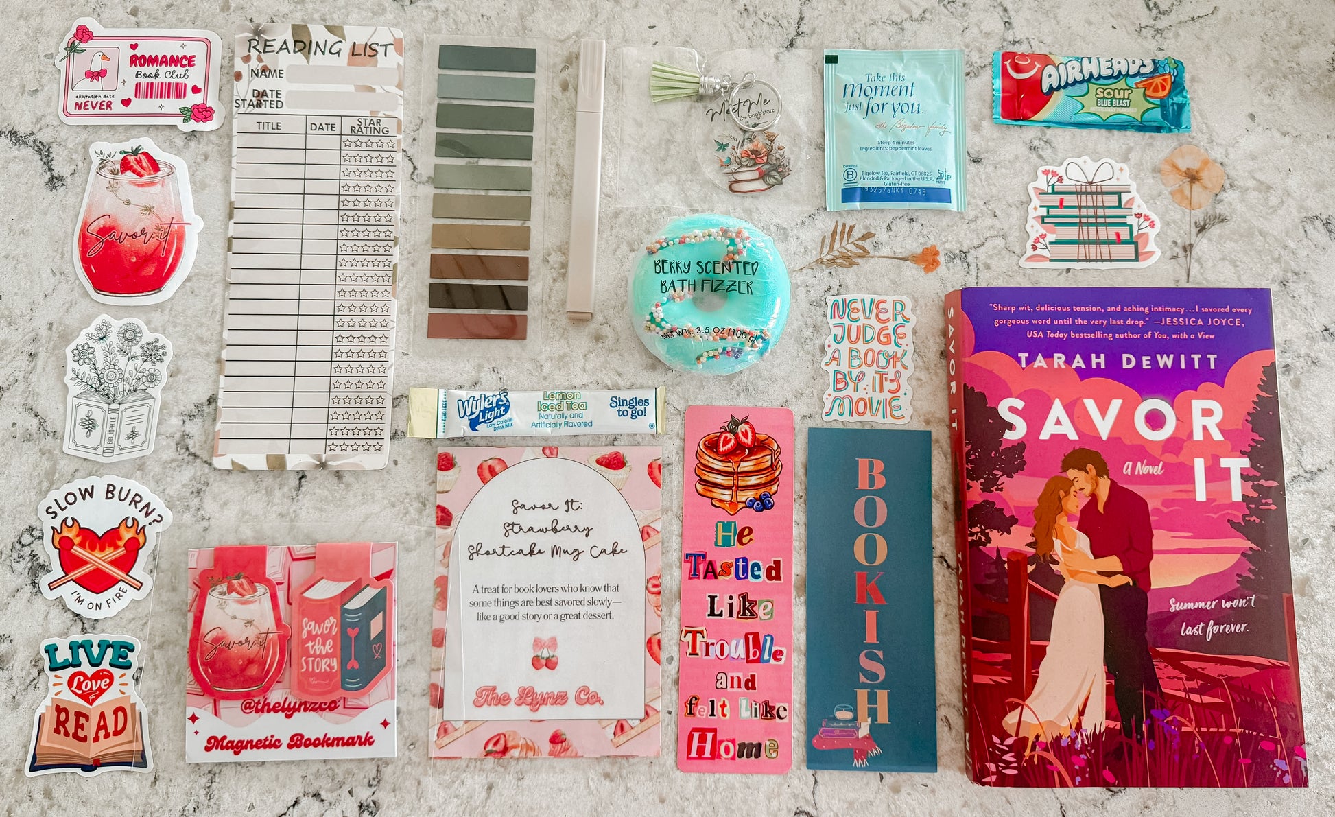 Flat lay of a romance-themed book box featuring the novel “Savor It” by Tarah DeWitt, surrounded by bookish extras including magnetic bookmarks, reading tracker, bath bomb, stickers, tea, candy, and a strawberry shortcake mug cake recipe card — styled on a marble background.