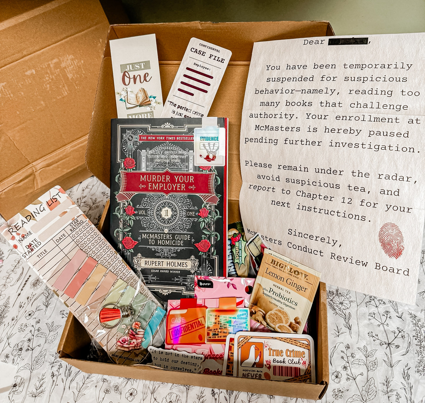 Open mystery-themed book box featuring “Murder Your Employer” by Rupert Holmes, surrounded by true crime extras including bookmarks, tea, reading list, stickers, magnetic tabs, keychain, and a fake suspension notice styled as an investigation letter — all inside a brown shipping box on a black-and-white floral sheet.