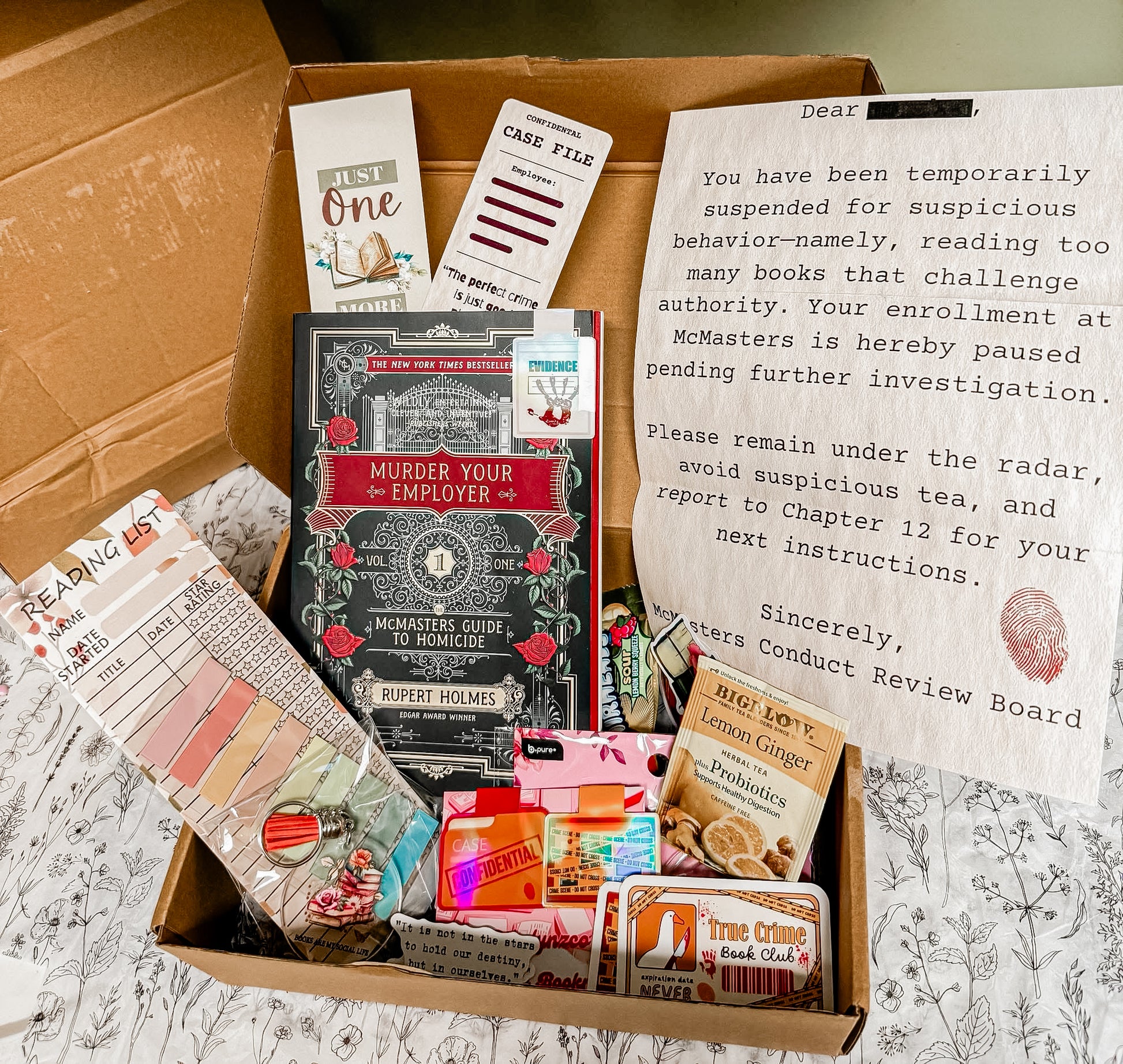 Open mystery-themed book box featuring “Murder Your Employer” by Rupert Holmes, surrounded by true crime extras including bookmarks, tea, reading list, stickers, magnetic tabs, keychain, and a fake suspension notice styled as an investigation letter — all inside a brown shipping box on a black-and-white floral sheet.